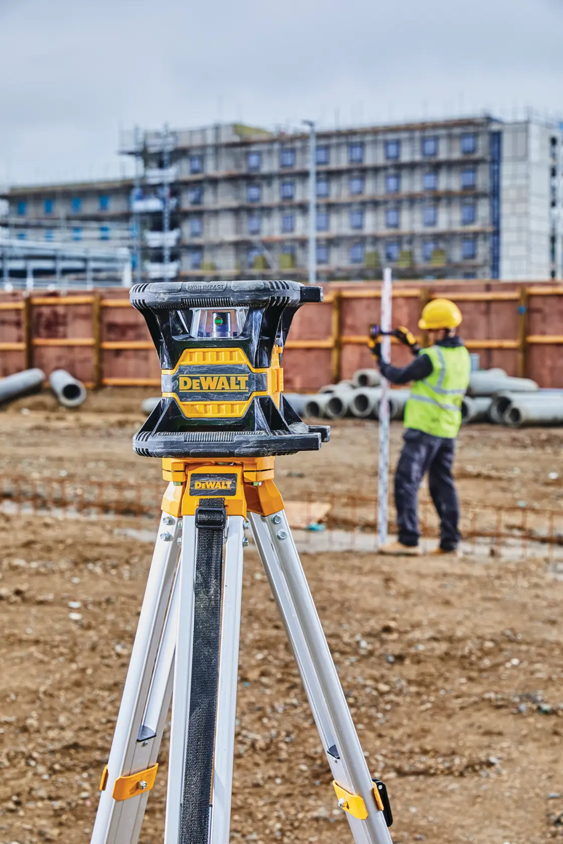 A DEWALT laser level mounted on a tripod at a construction site, with a worker wearing a yellow hard hat and safety vest using a measuring rod in the background. Pipes and a building under construction are also visible.