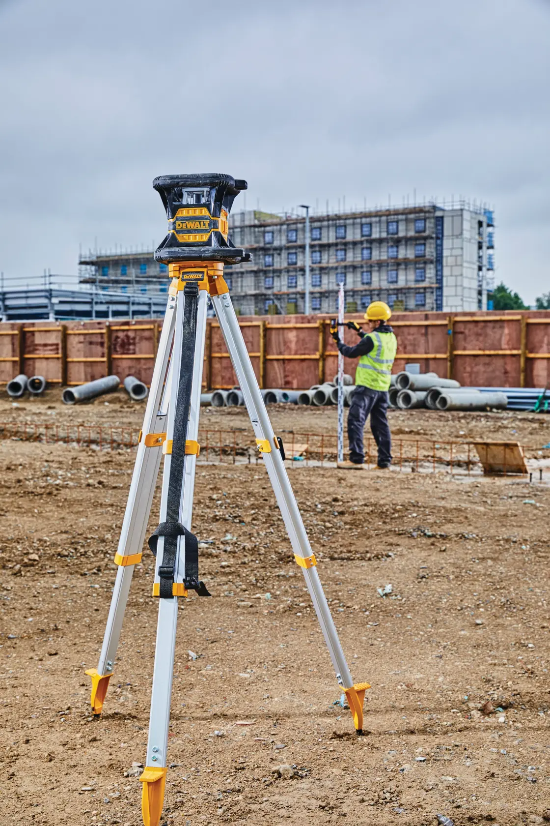A DEWALT DW080LRS laser level mounted on a tripod at a construction site, with a worker in a safety vest and helmet measuring in the background.