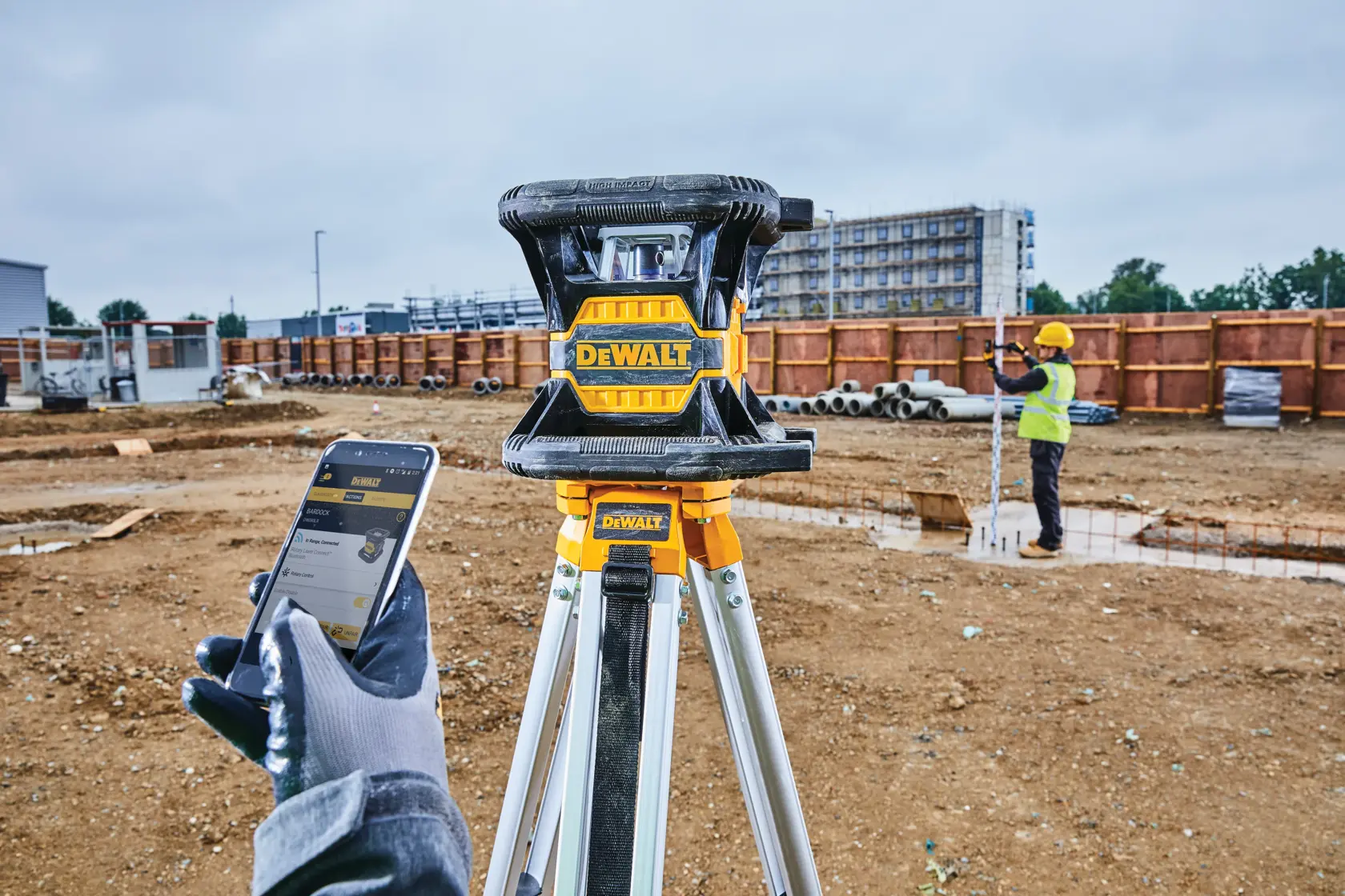 A DEWALT DW080LRS laser level positioned on a tripod at a construction site, with a worker in safety gear standing in the background near pipes and a partially built structure.