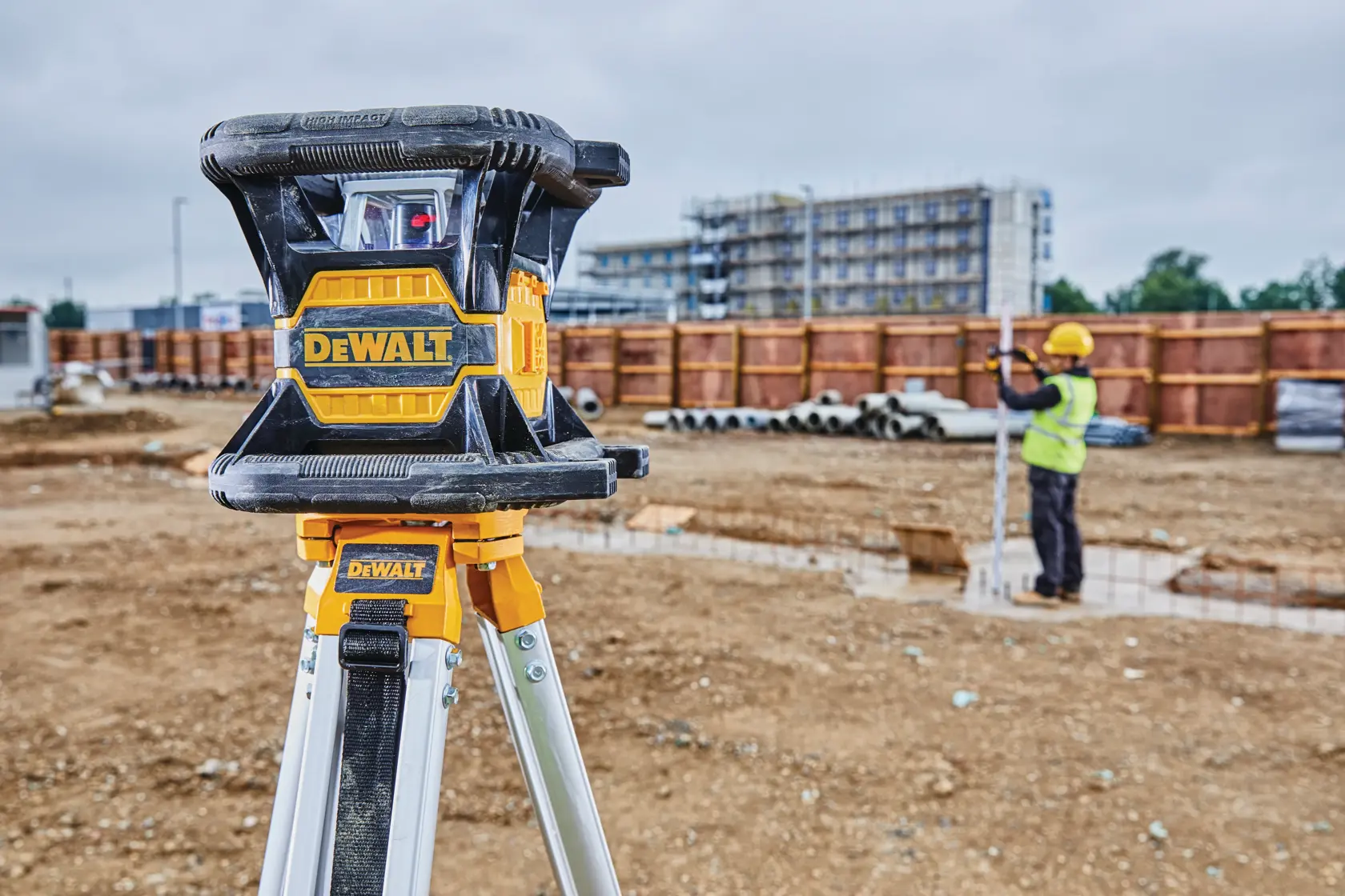 A DEWALT laser level mounted on a tripod at a construction site. A person wearing gloves is holding a smartphone near the device, and another worker in a safety vest and helmet is visible in the background.