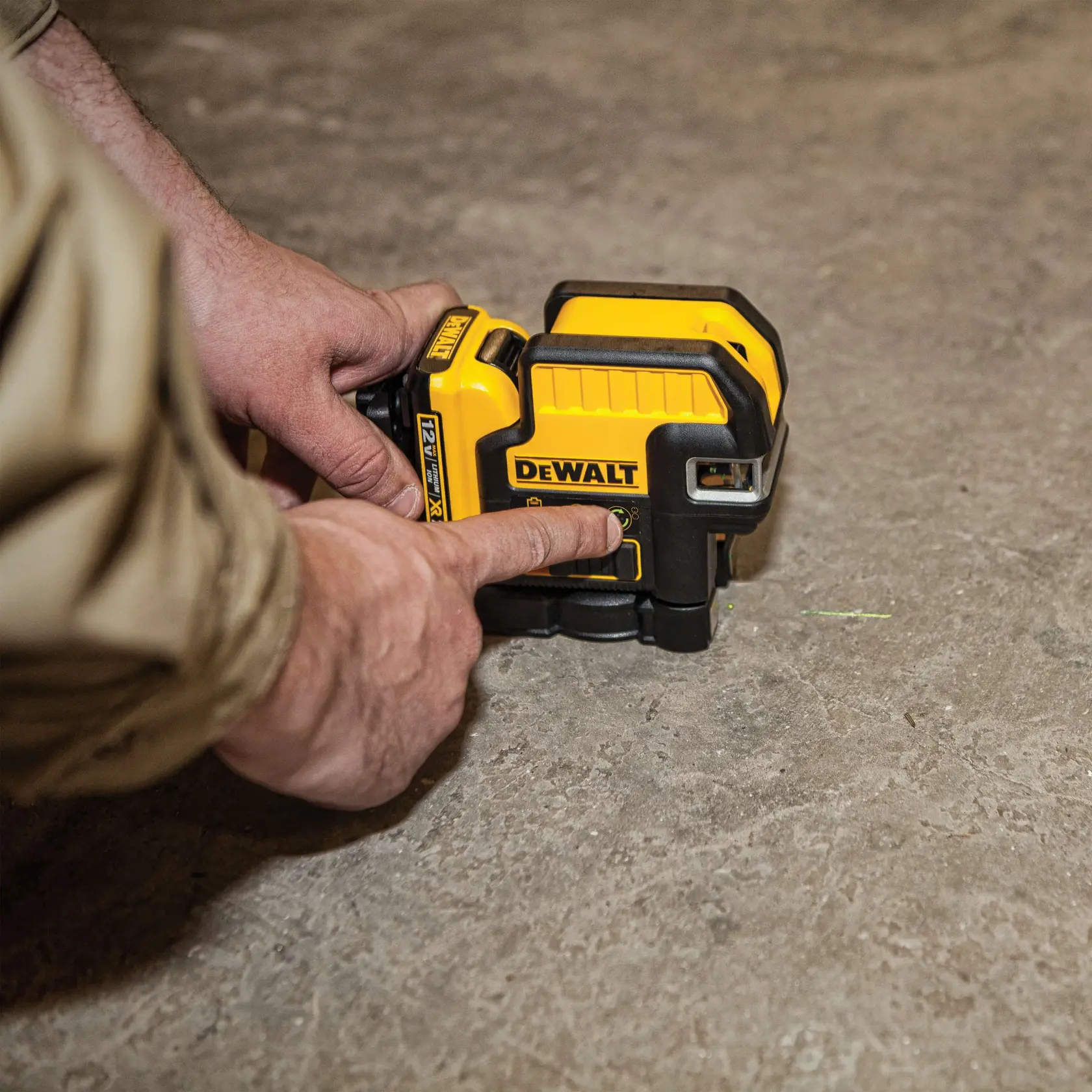 A person is operating a yellow and black DEWALT laser level tool placed on a concrete floor.