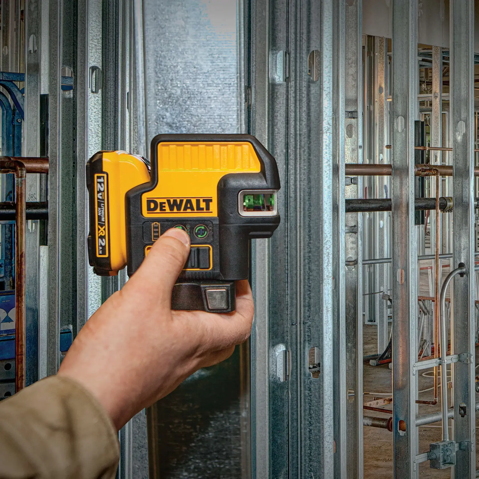A hand holding a yellow and black DEWALT laser level device, model DW0822LG, against metal wall studs at a construction site.