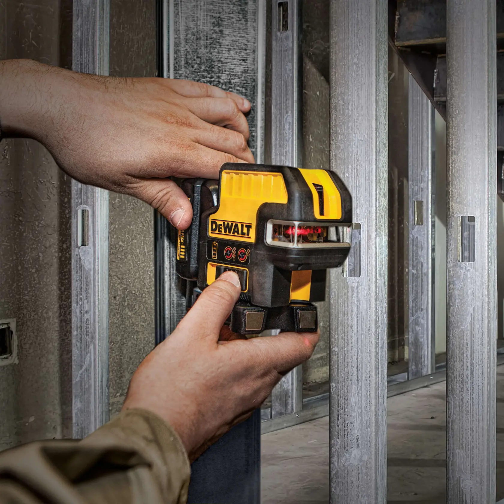 A person uses a DEWALT DW0822LR laser level tool to measure and align metal studs on a construction site. The tool emits a red laser line and is held against the metal framing.