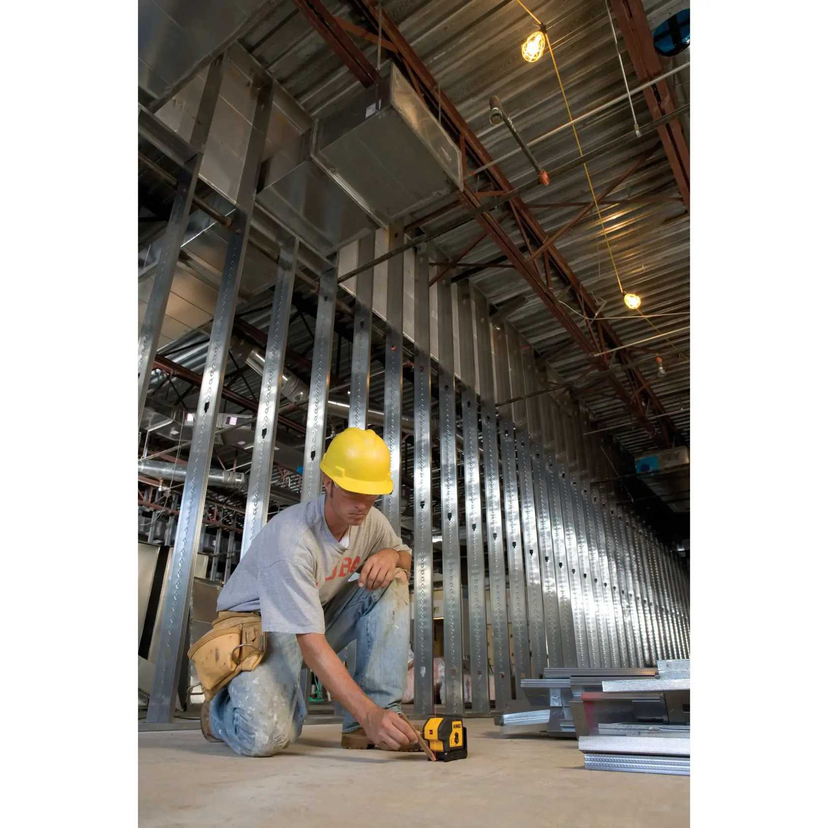 A construction worker wearing a yellow hard hat is kneeling on the floor of a building under construction. The worker is using a yellow and black DEWALT laser level tool (model DW083K) on the concrete floor, surrounded by metal wall studs and construction materials.