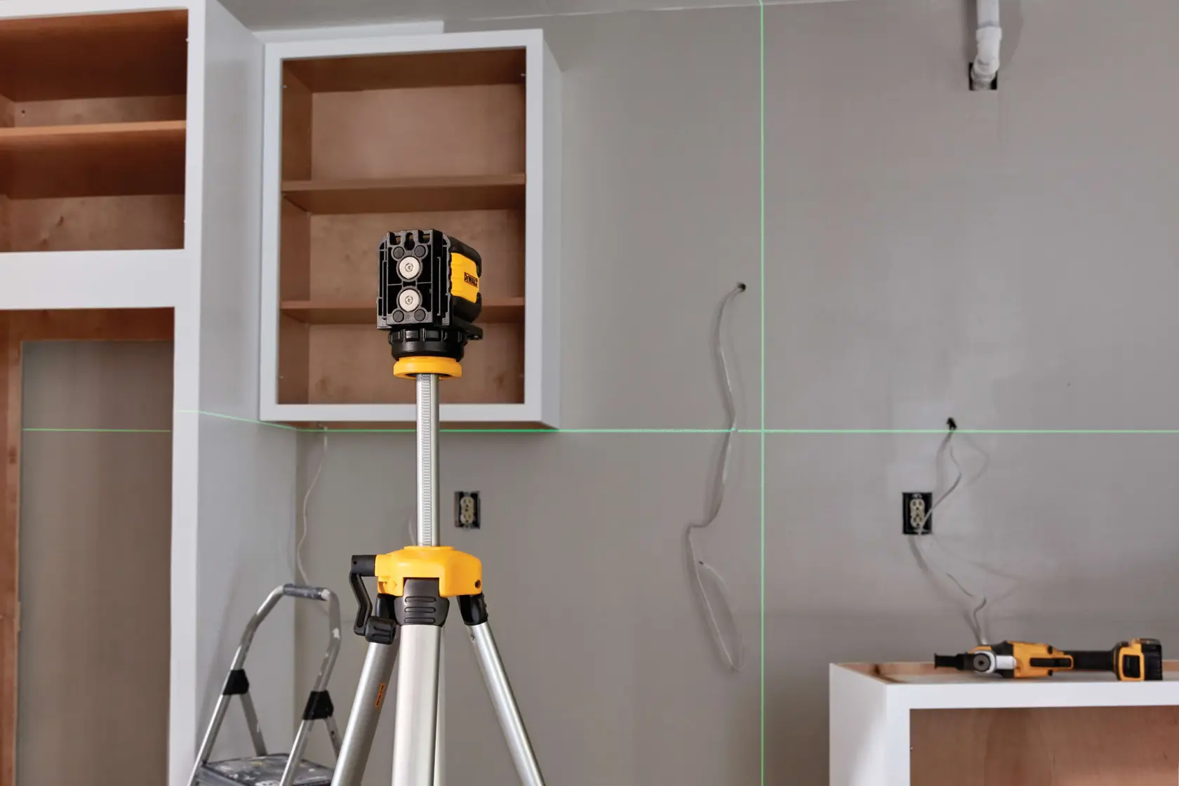 A DEWALT laser level device mounted on a tripod is positioned in a kitchen under construction. Green laser lines are projected horizontally and vertically onto the walls. A step ladder, electrical wires, outlets, and power tools are visible in the background.