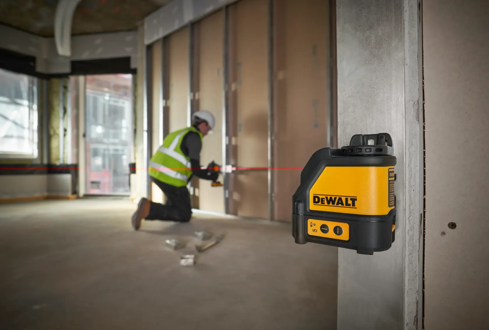 A DEWALT DW088K cross line laser level is mounted on a metal stud in a construction site. The laser device is emitting red lines for precise alignment, and construction materials are visible in the background.