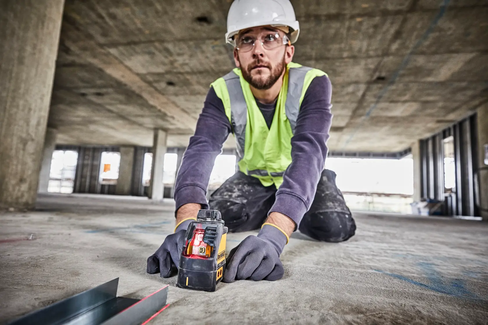 Worker using a DEWALT DW088K laser level on a construction site floor, wearing gloves and safety gear.