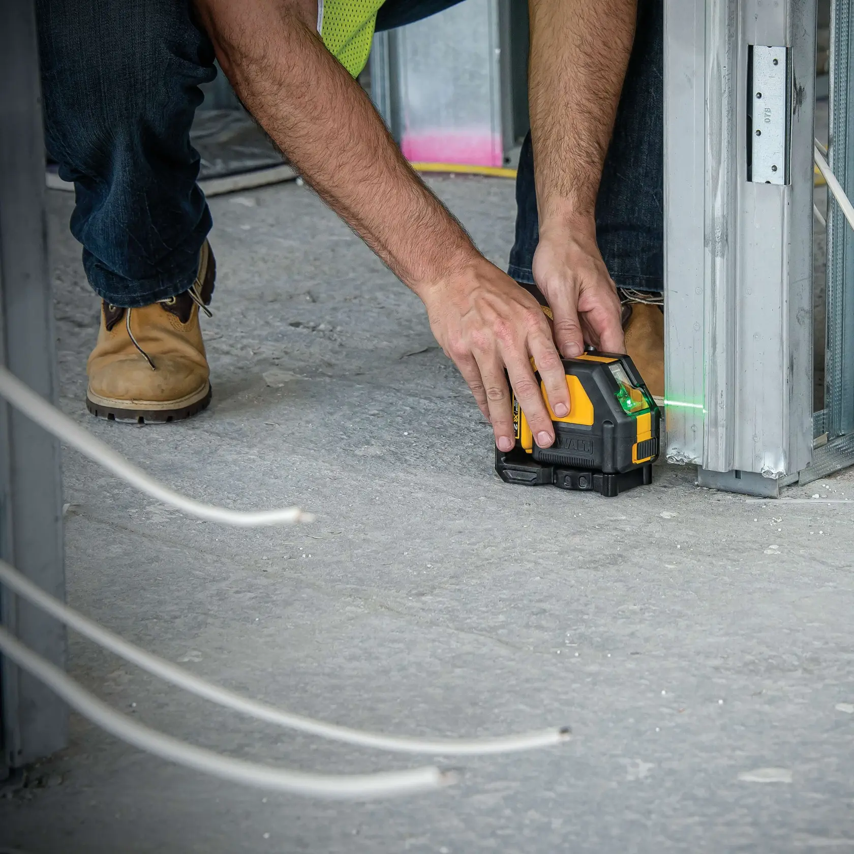 A person is placing a DEWALT DW088LG green beam laser level on a construction site floor, aligning its laser with metal framing for accurate measurements.