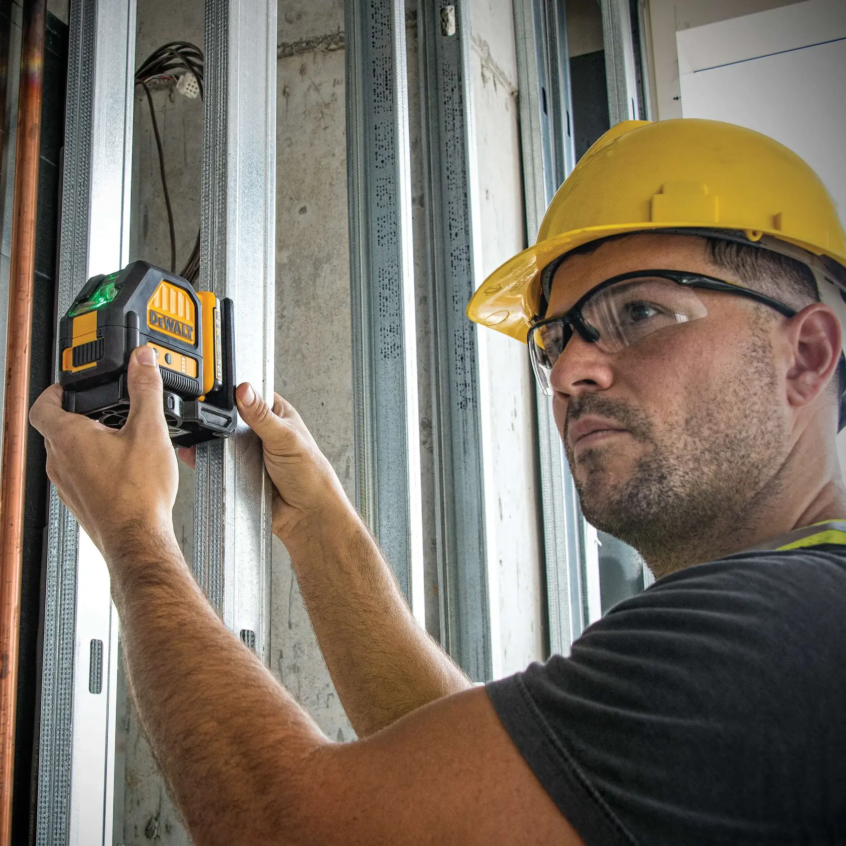 A person wearing a yellow hard hat uses a DEWALT DW088LG green laser level against a metal frame at a construction site. The person's face is blurred.