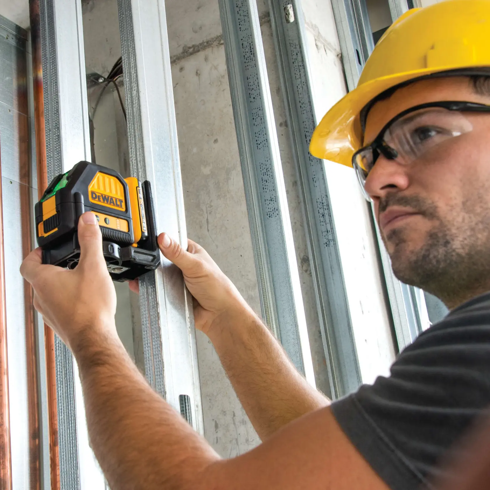A person wearing a yellow safety helmet is using a DEWALT laser level tool to align metal framing studs at a construction site.