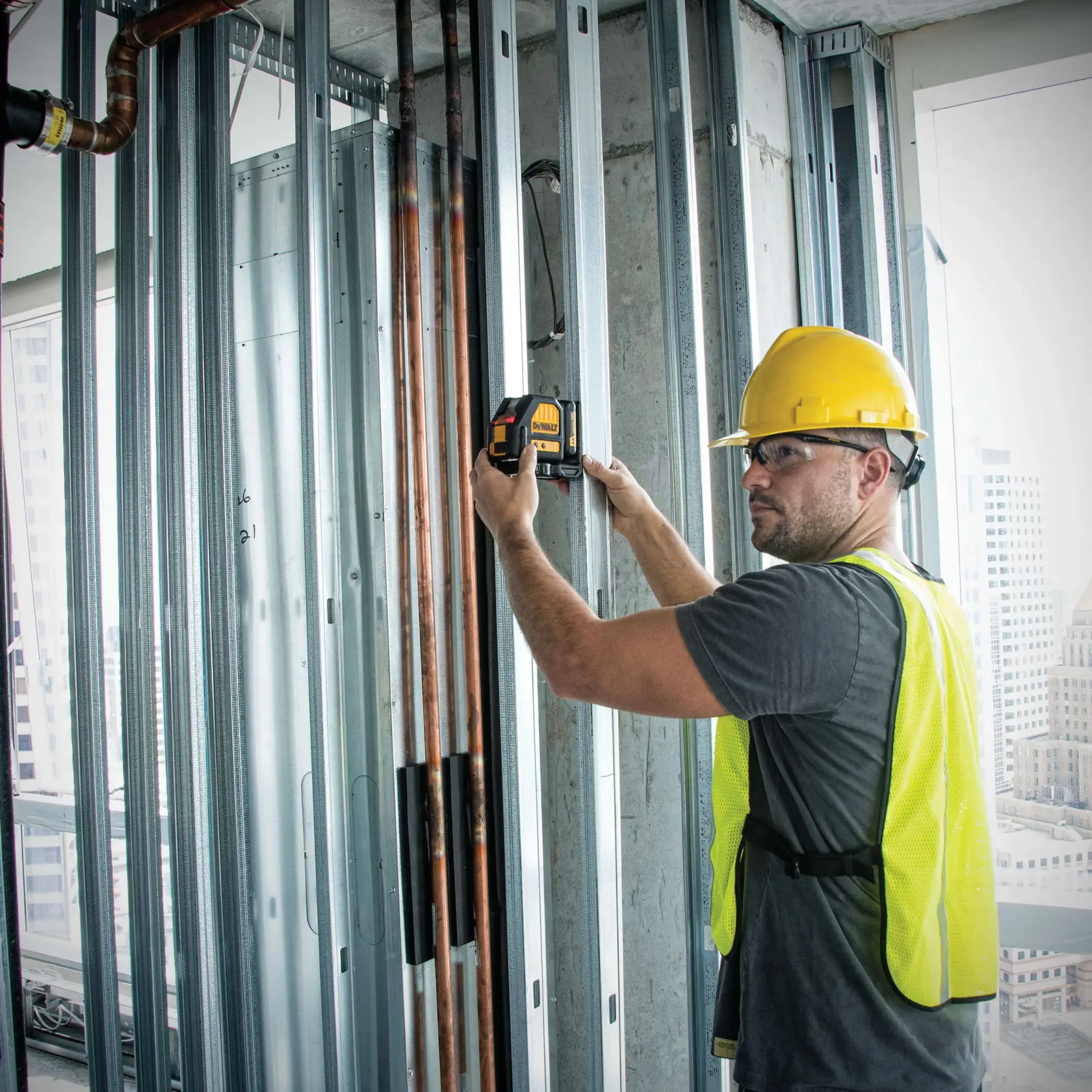 A worker wearing a yellow hard hat and safety vest is using a DEWALT laser level on a metal stud wall in a construction site.