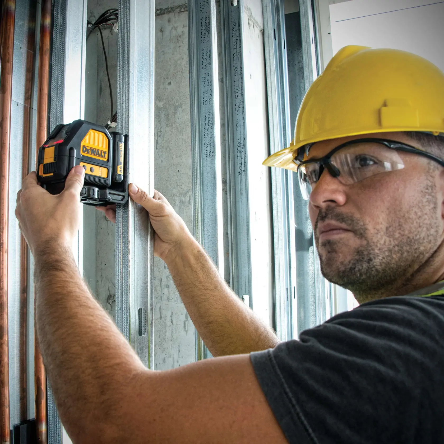 A person wearing a yellow hard hat uses a DEWALT DW088LR laser level on a construction site, positioning it against metal wall framing.