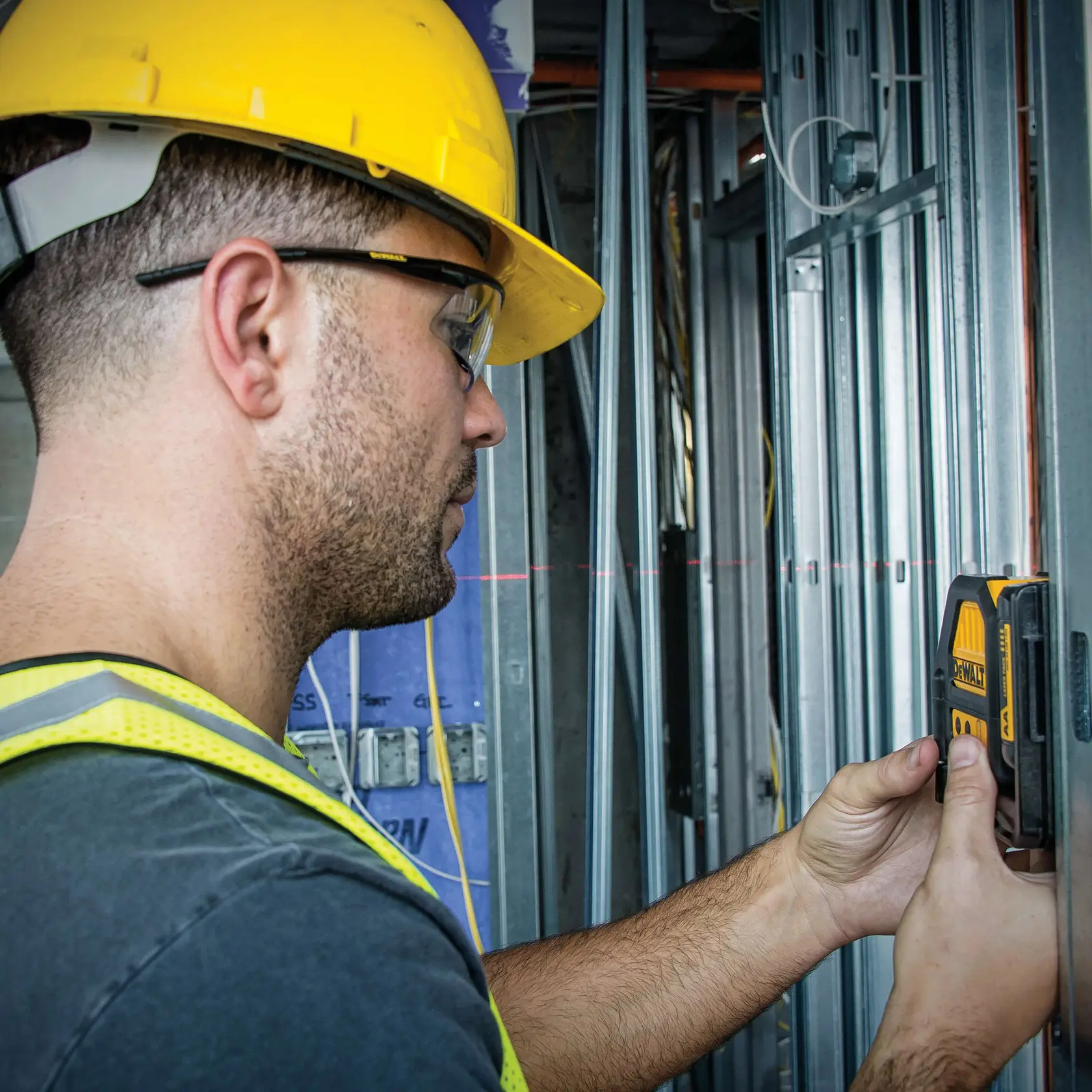 A person wearing a yellow hard hat and safety vest uses a DEWALT DW088LR laser level against a metal frame on a construction site.