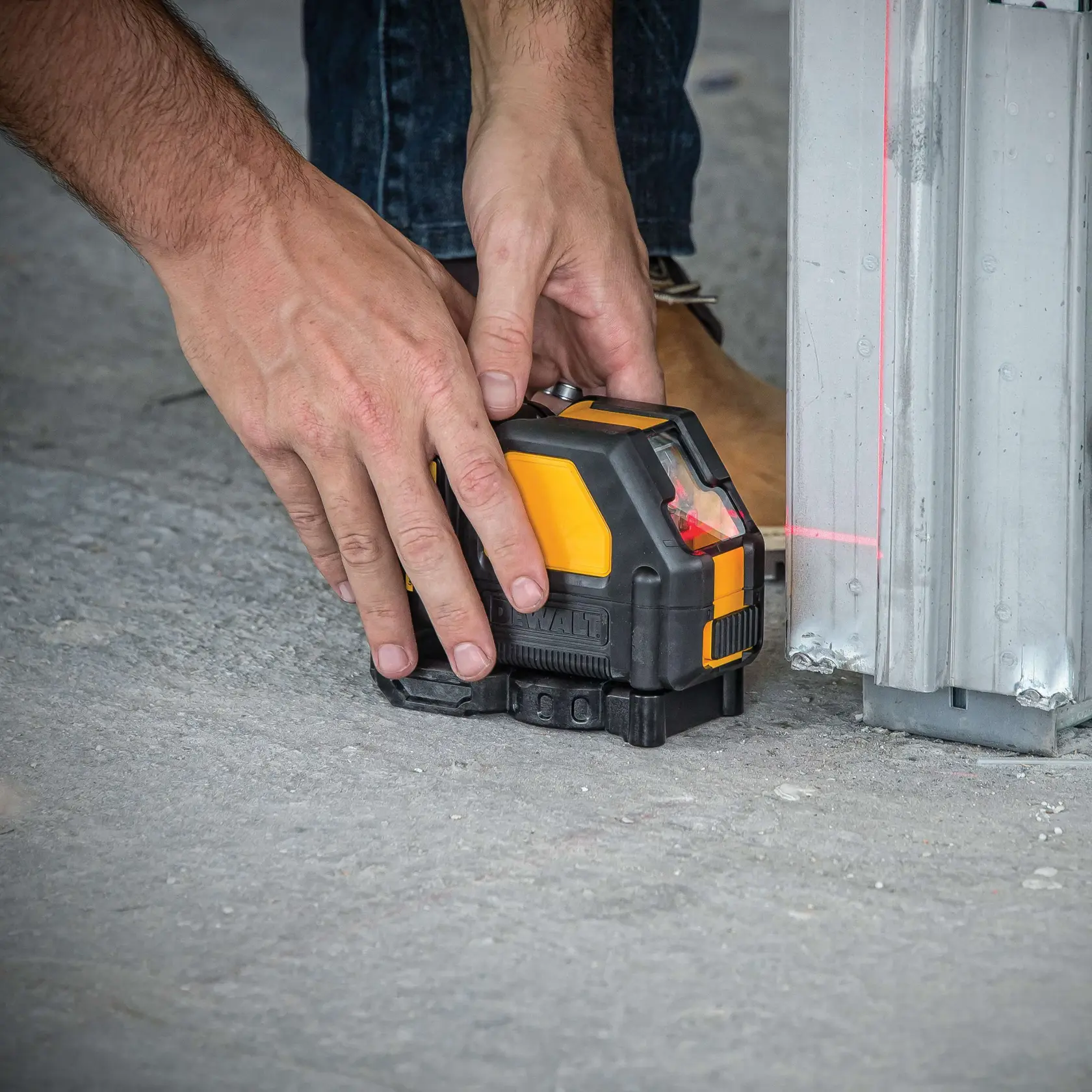 A person is positioning a DEWALT DW088LR laser level on a concrete floor next to a metal structure, with visible red laser lines for alignment.