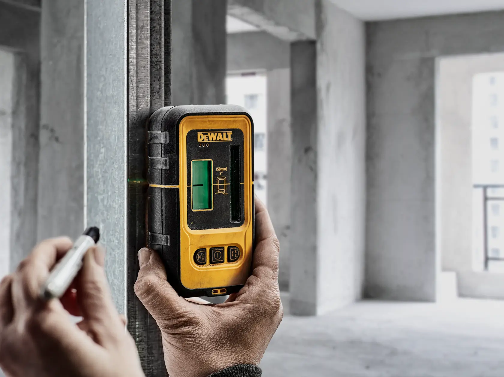 A person uses a DEWALT DW0892G green laser detector attached to a metal beam in a construction site, marking a line with a marker.