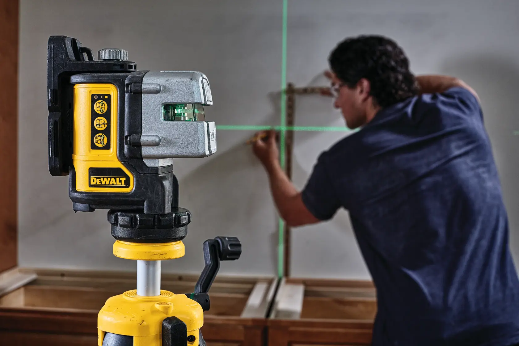 A DEWALT DW089CG green line laser level is mounted on a tripod in the foreground, projecting horizontal and vertical green laser lines onto a wall. In the background, a person is marking the wall using the laser guides for precision alignment.