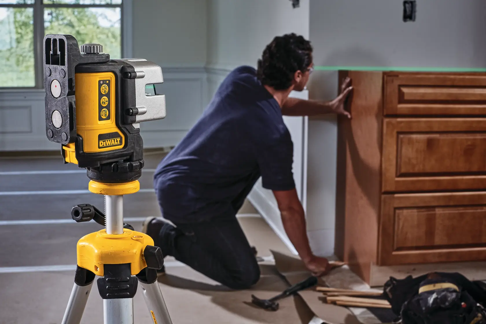 A DEWALT DW089CG green beam laser level mounted on a tripod in the foreground, with a person installing cabinetry in the background in a residential room.