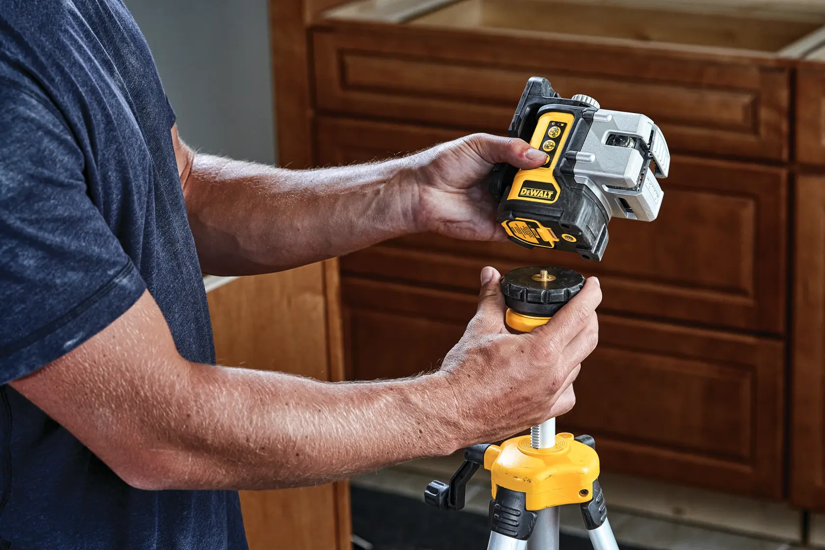 A person is mounting a DEWALT DW089CG green cross line laser on a yellow tripod in a home setting with wooden cabinets in the background.