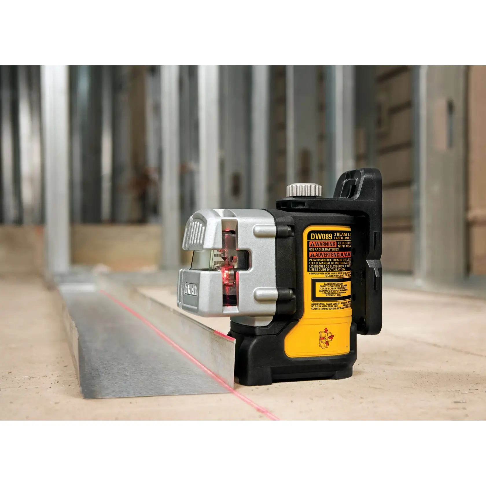 DEWALT DW089K three-beam laser level placed on a construction site floor, projecting a red laser line along a metal framing track with metal wall studs in the background.