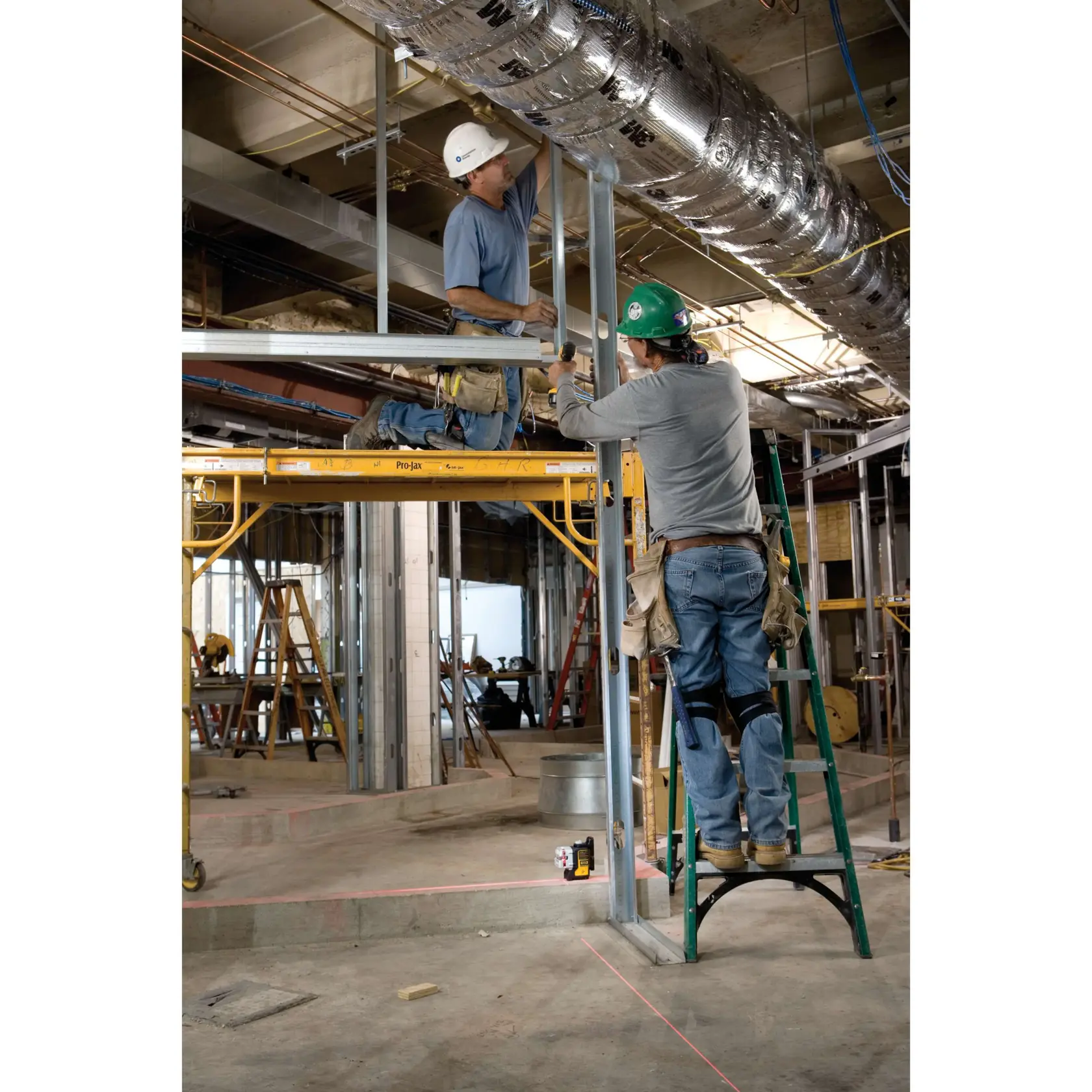 Two construction workers are assembling metal framing in an indoor construction site. One worker stands on a green ladder, while the other kneels on yellow scaffolding. A DEWALT DW089K laser level is placed on the floor below, projecting red laser lines to assist with alignment. The workspace is filled with construction equipment, ductwork, and scaffolding.