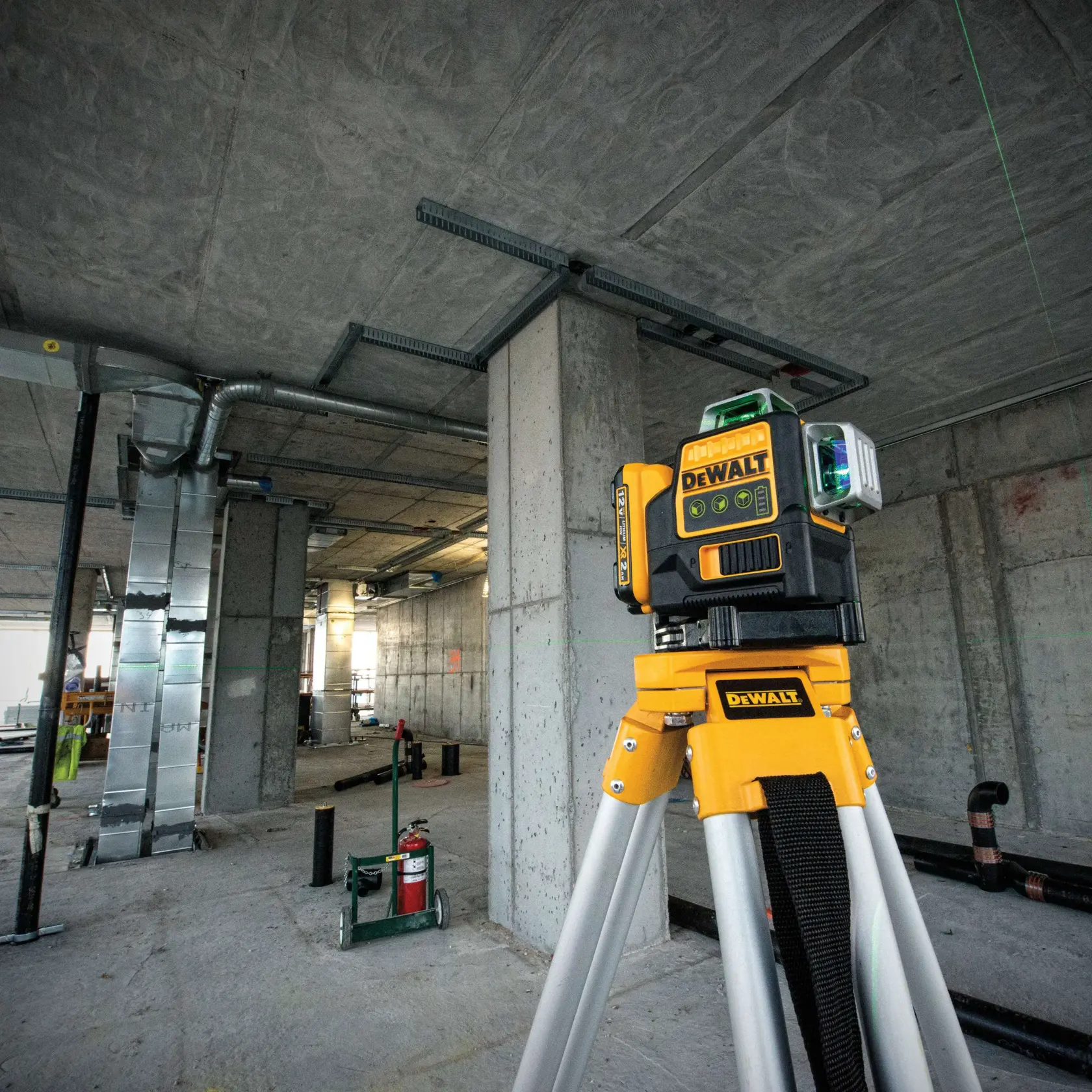 DEWALT DW089LG green beam laser level mounted on a tripod in a concrete construction site, surrounded by industrial equipment and unfinished walls.