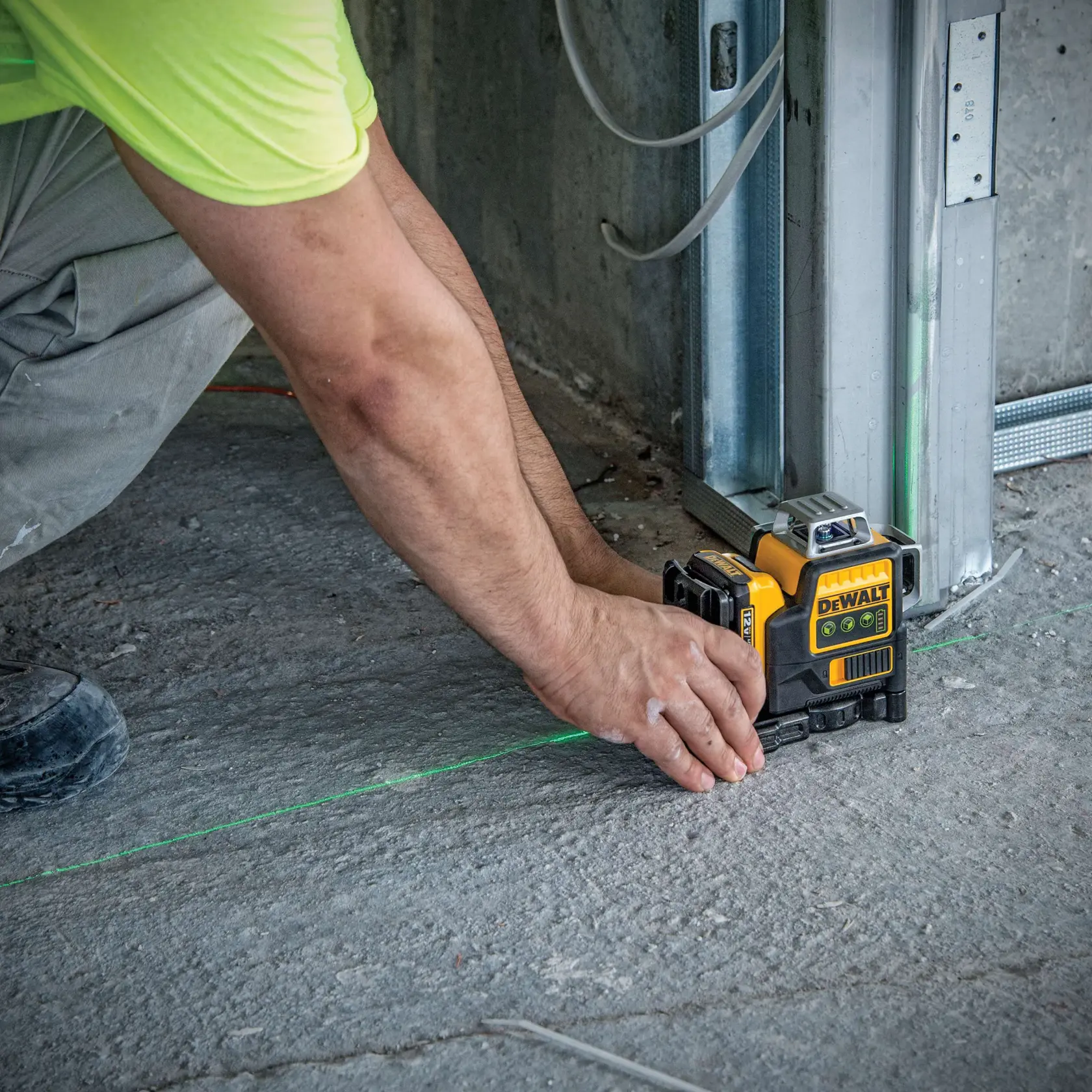 A person using a DEWALT laser level (model DW089LG) on a construction site floor to project green laser lines for accurate alignment near a metal frame.