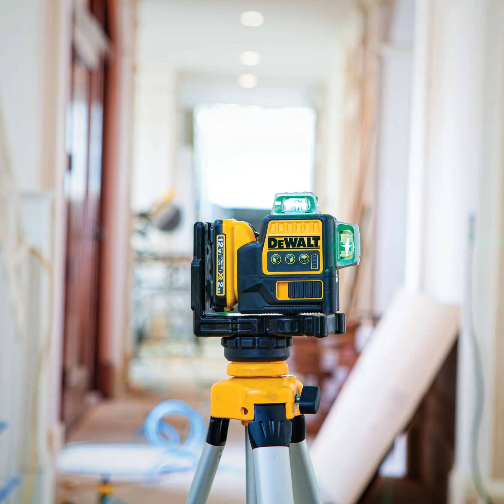A DEWALT DW089LG green beam line laser level is mounted on a tripod in an indoor construction or renovation setting, with blurred background showing tools and materials.