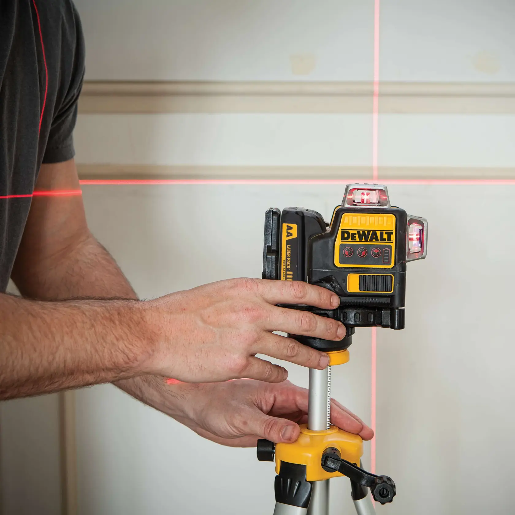 A person adjusts a DEWALT DW089LR laser level mounted on a tripod. The device projects visible horizontal and vertical red laser lines onto a wall for precise leveling and alignment.