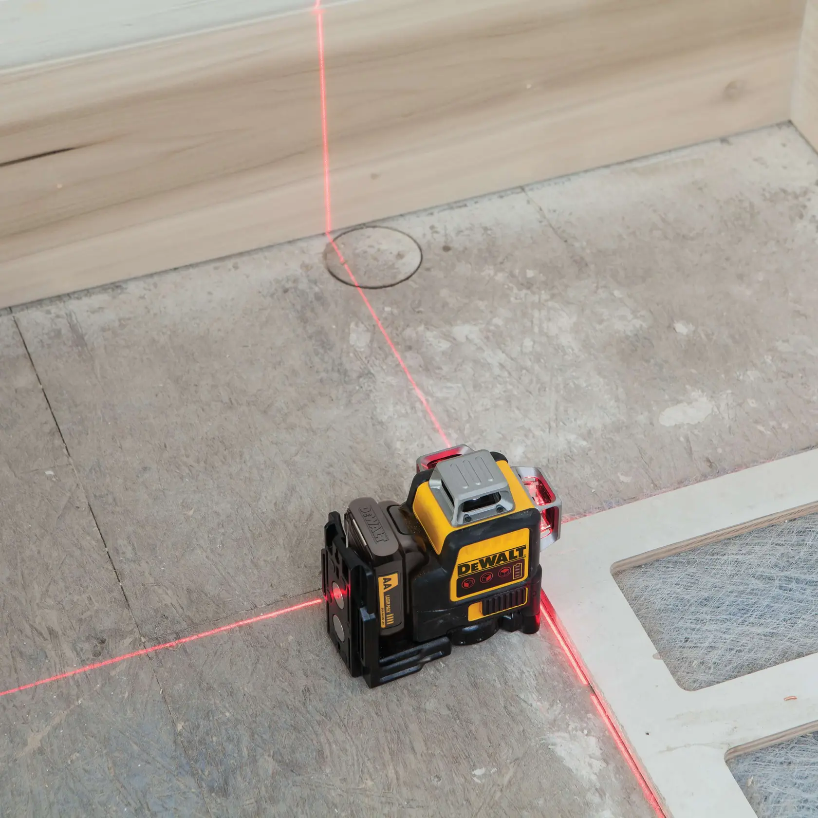 A DEWALT DW089LR red laser level tool is placed on a construction site floor, projecting vertical and horizontal red laser lines for alignment.