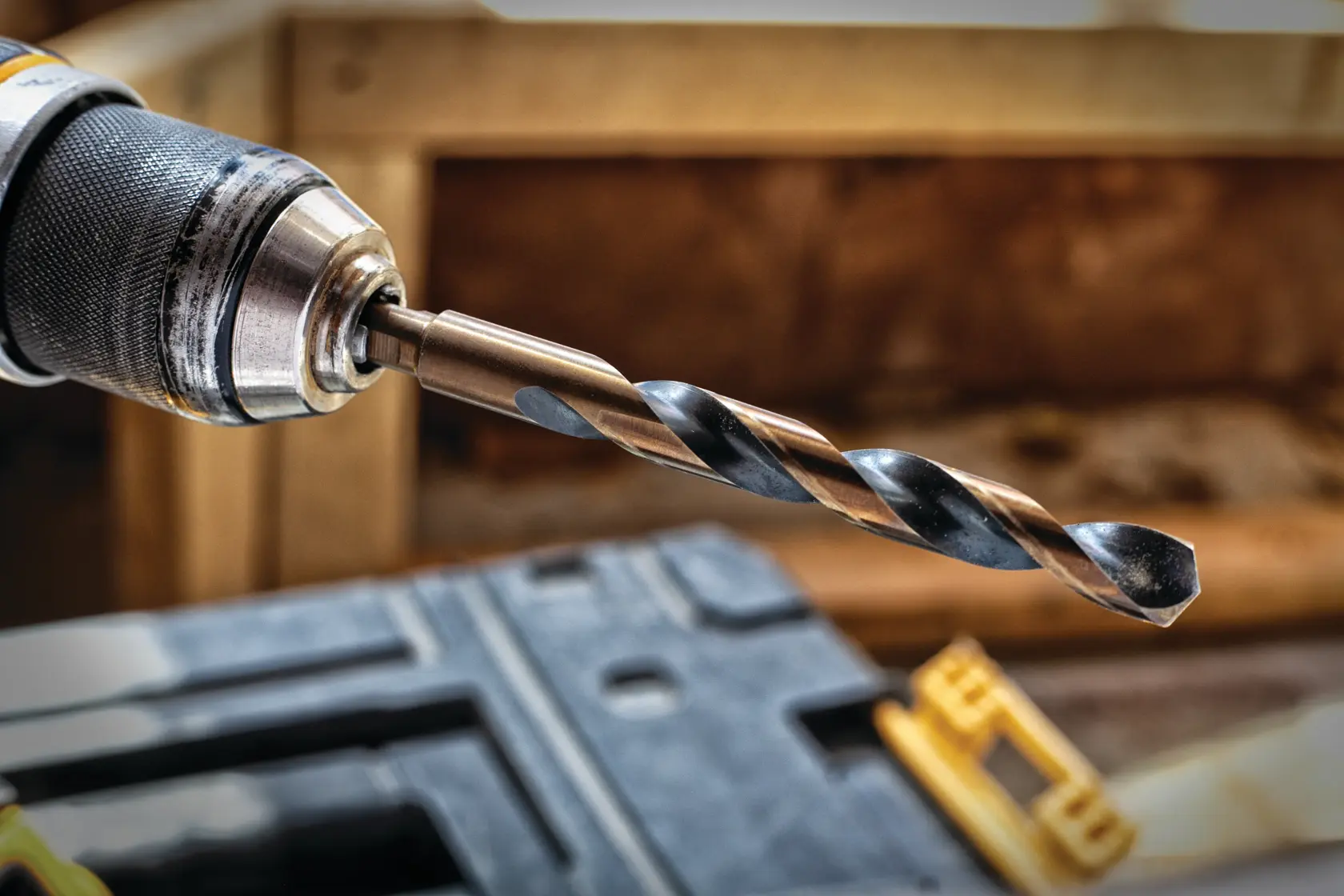 Close-up view of a drill with a metal twist drill bit attached, shown in a workshop setting with tools and wood in the background.