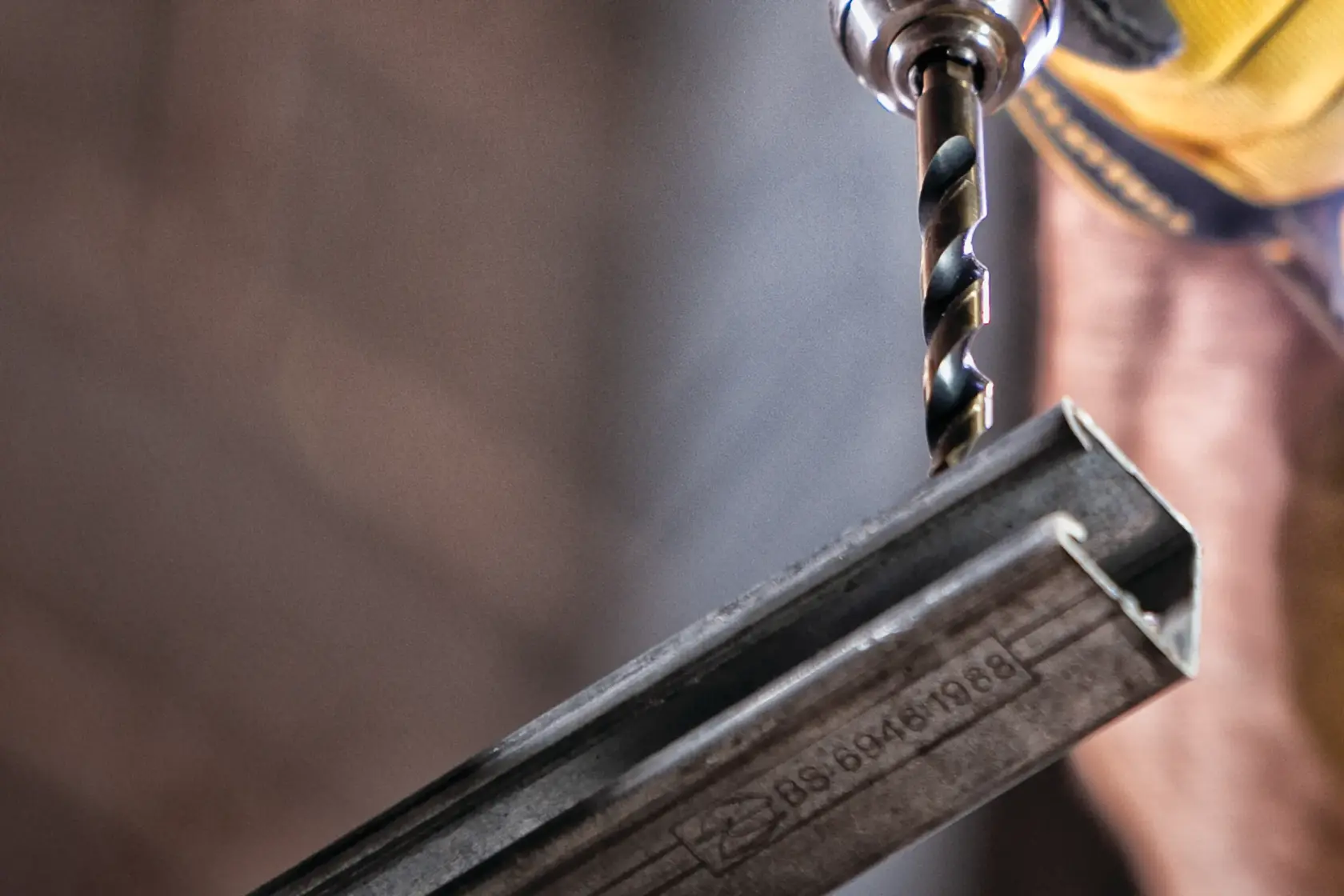 A close-up of a DEWALT drill bit being used to drill into a metal angle bar, held securely in place. The focus is on the drill bit and the bar, with a blurred background.