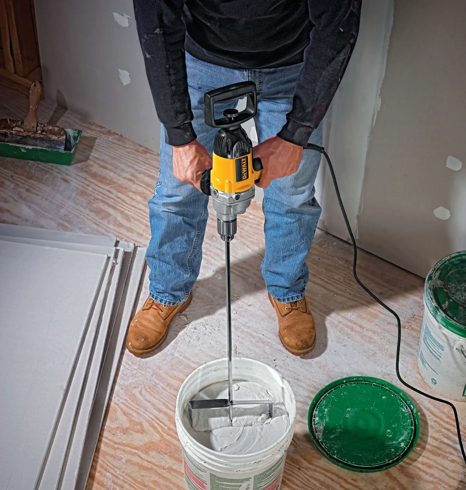 A person using a DEWALT DW130V electric drill, equipped with a mixing paddle, to stir white compound inside a bucket. The scene is set on a wooden floor with sheets of drywall and other construction materials visible.