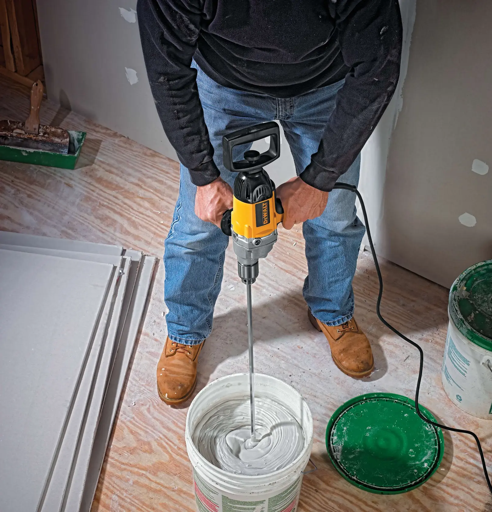 A person using a DEWALT DW130V power drill to mix plaster or joint compound in a large bucket, with drywall panels and tools visible on the floor.