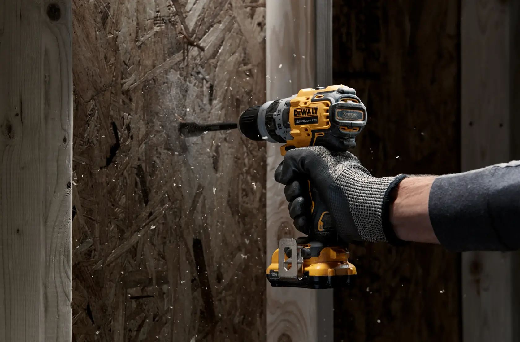 A person wearing a glove is using a DEWALT cordless power drill to drill into a wooden board, with wood debris flying around.