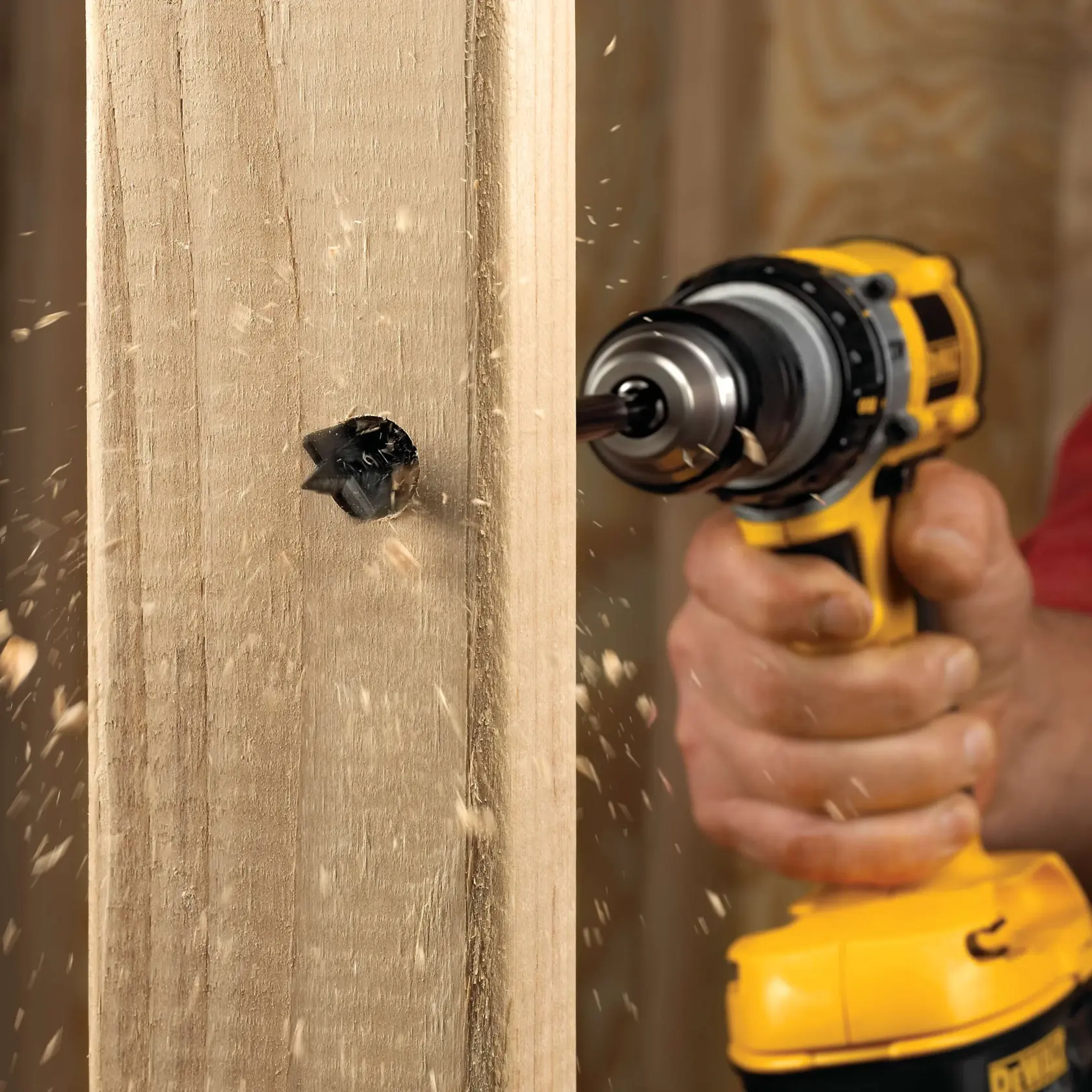 A close-up image of a person using a yellow DEWALT drill to create a hole in a wooden plank, with wood shavings flying as the drill bit penetrates the surface.
