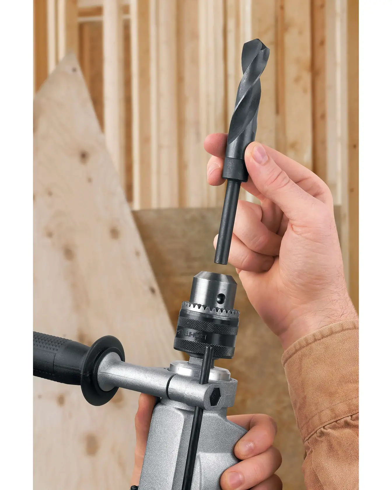 A person is inserting a large drill bit into the chuck of a power drill. The scene appears to take place in a construction setting, with wooden beams and plywood panels in the background.