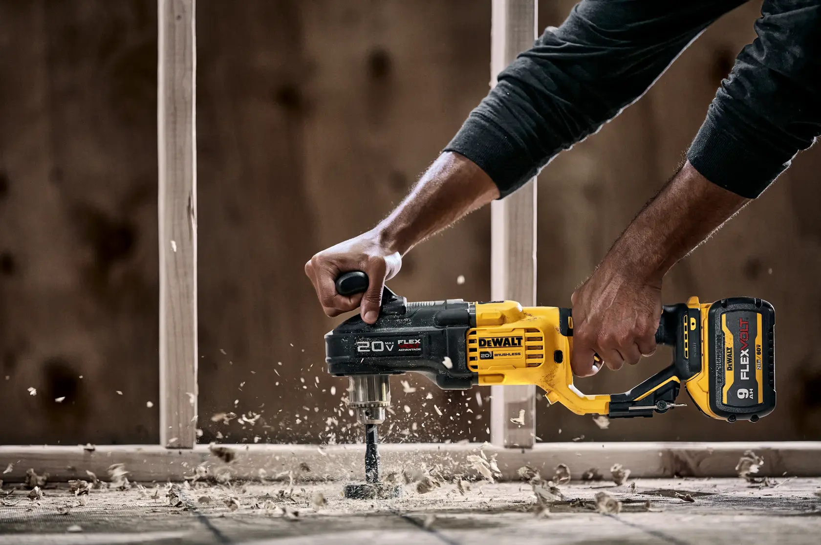 A person uses a DEWALT 20V FLEX cordless drill to bore a hole in wooden flooring, with wood shavings flying around. The drill has a yellow and black design and a FLEXVOLT battery attached.