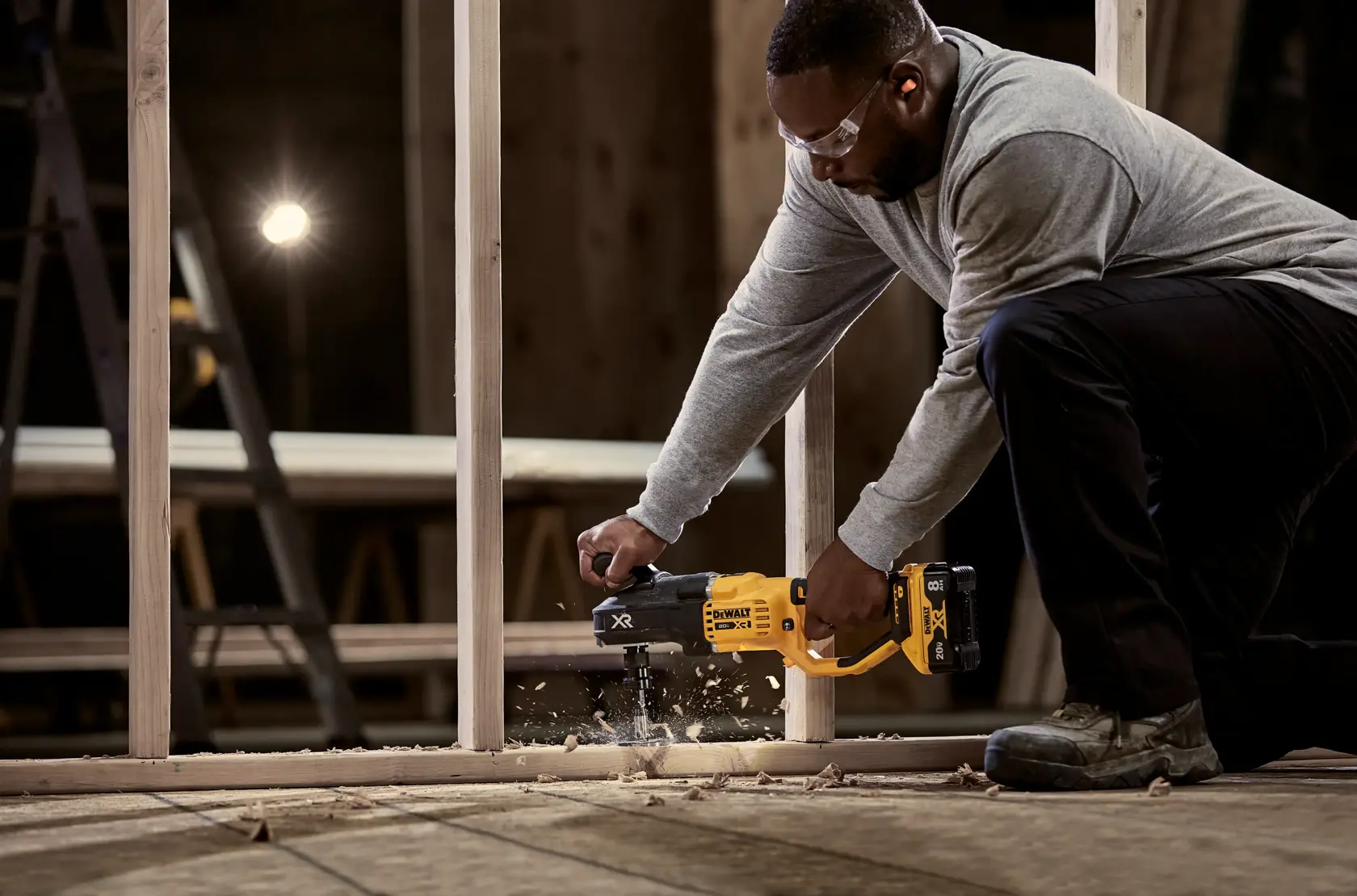 A person kneeling and using a DEWALT cordless drill (model DW1639) to drill through wood framing at a construction site. Wood shavings are seen flying from the drilled hole, and the background includes a ladder and work light.