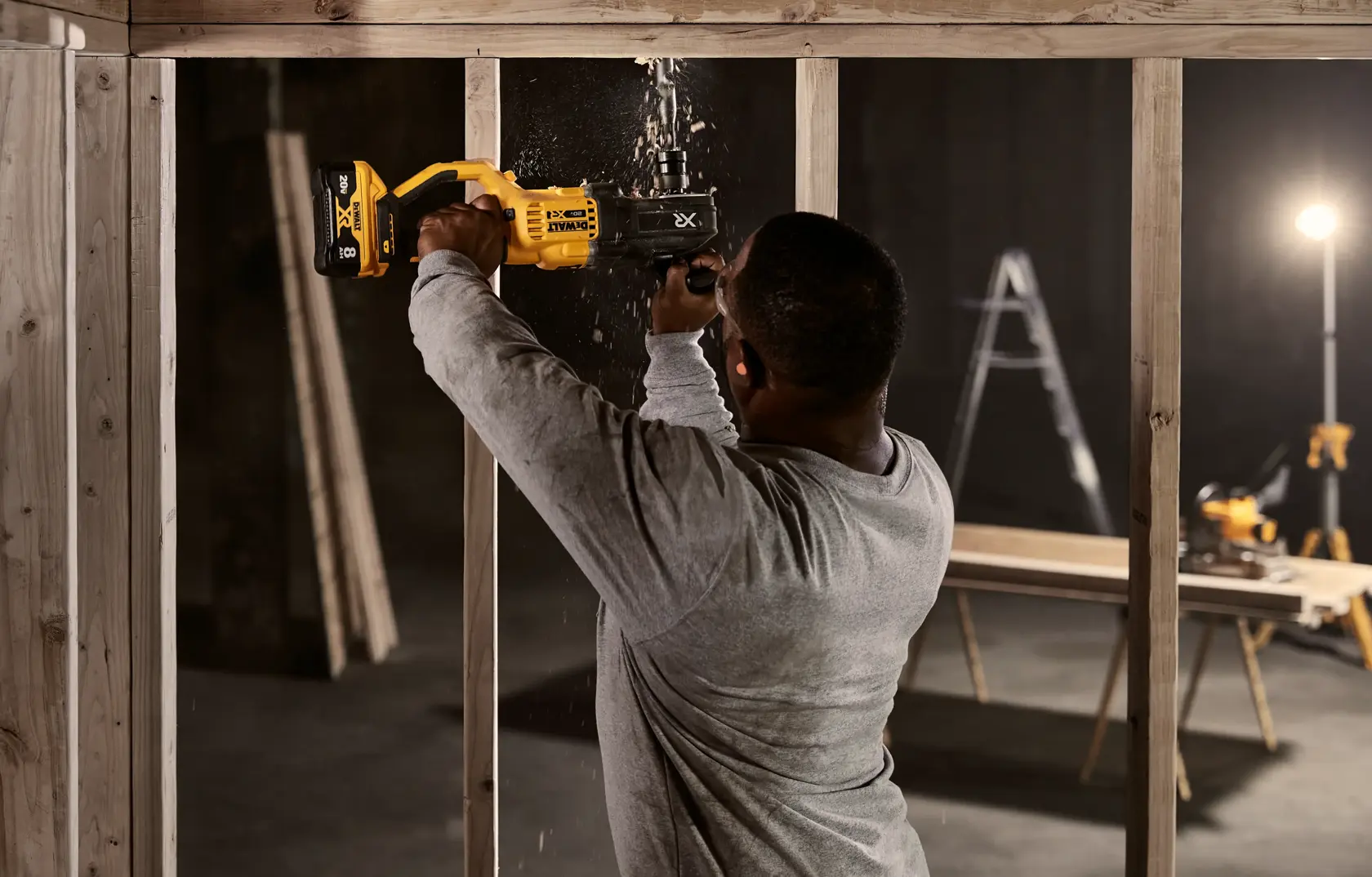 A person using a yellow DEWALT power tool to cut through wooden framing in a construction setting. Sawdust is flying as the tool operates. There is a ladder, a workbench, and lighting equipment in the background.