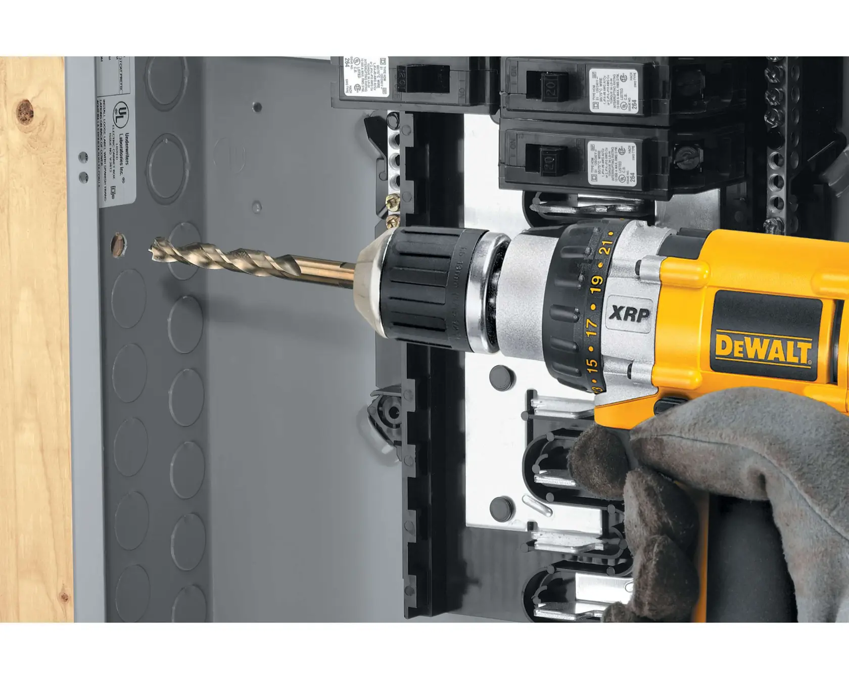 A DEWALT cordless drill with a drill bit is being used to drill into a metal electrical panel. The drill is held by a gloved hand.