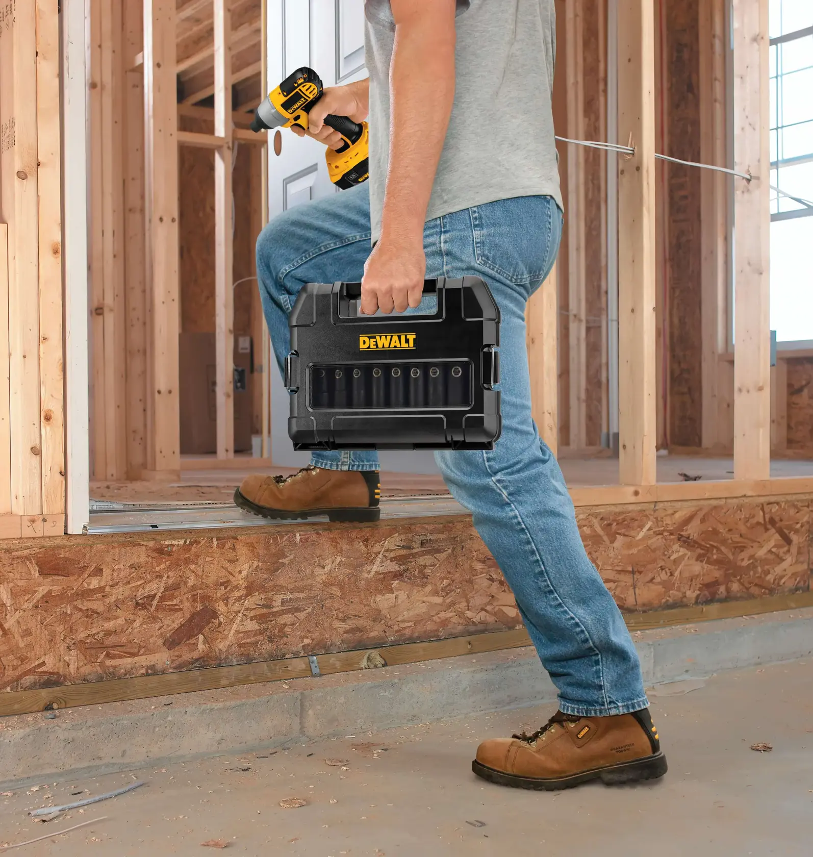A person in a construction area holding a DEWALT black tool case and a cordless drill, wearing jeans, boots, and a grey t-shirt.