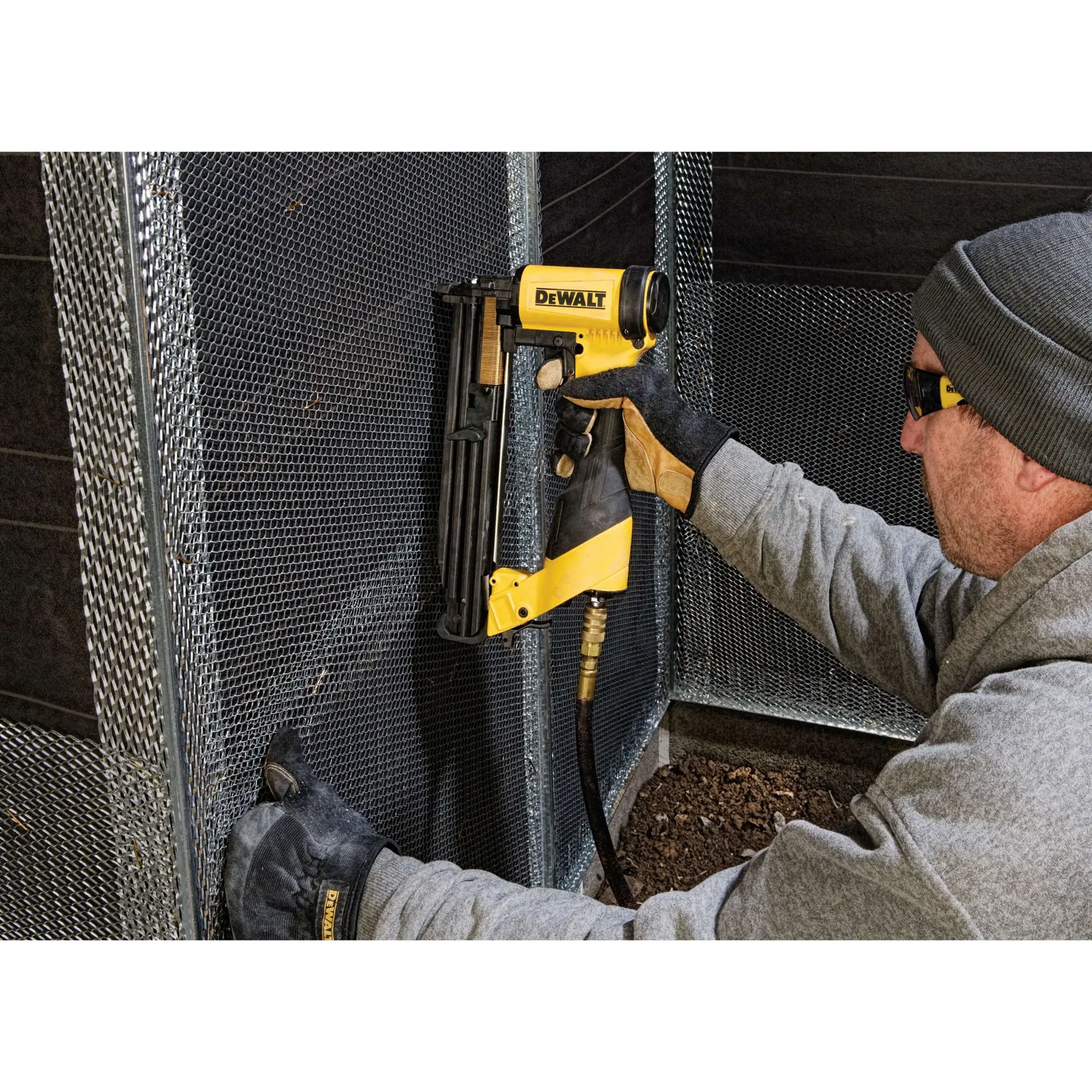 A person wearing gloves and a hoodie uses a DEWALT pneumatic nail gun to secure wire mesh onto a metal frame.