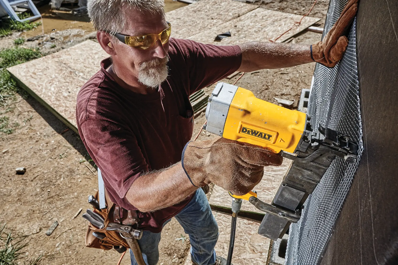 A person using a DEWALT pneumatic tool to attach metal mesh to an outdoor wall at a construction site. The individual is wearing gloves, a tool belt, and a maroon shirt.