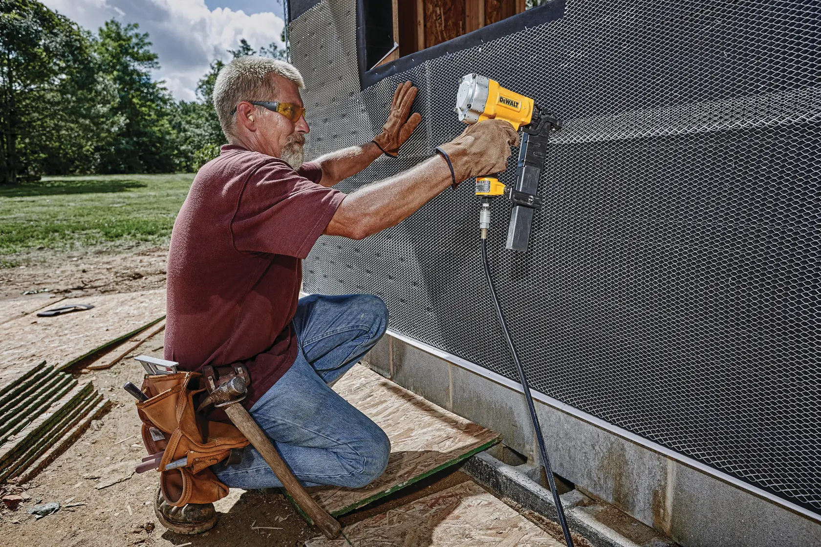 A person using a DEWALT pneumatic nail gun to fasten metal mesh on the exterior wall of a building under construction. The person is kneeling, wearing work gloves and a tool belt, with trees and grass visible in the background.