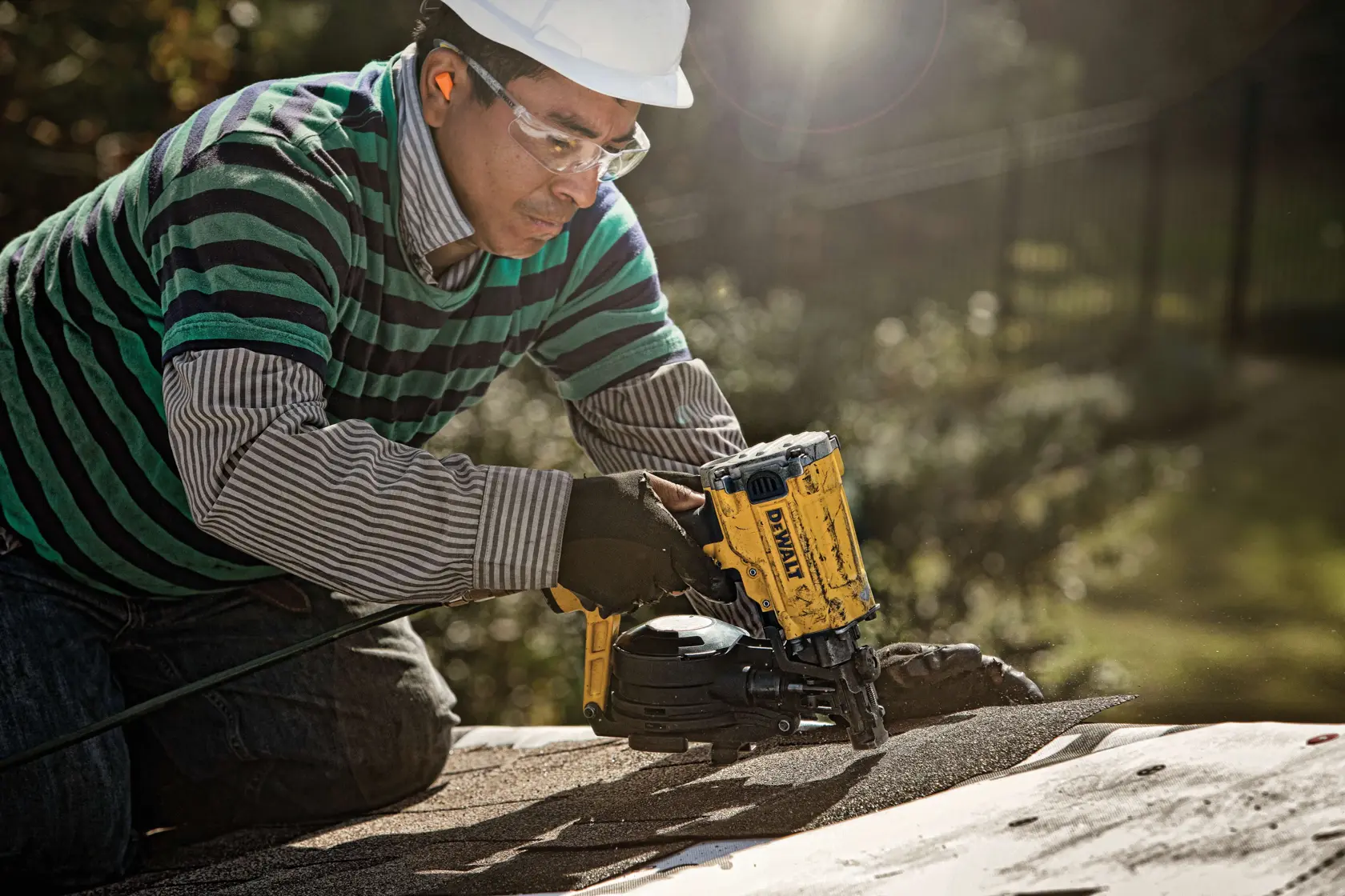 A person wearing a striped shirt and gloves uses a DEWALT DW45RN roofing nail gun on a roof surface outdoors. The individual's face is blurred for privacy and the setting is in daylight.