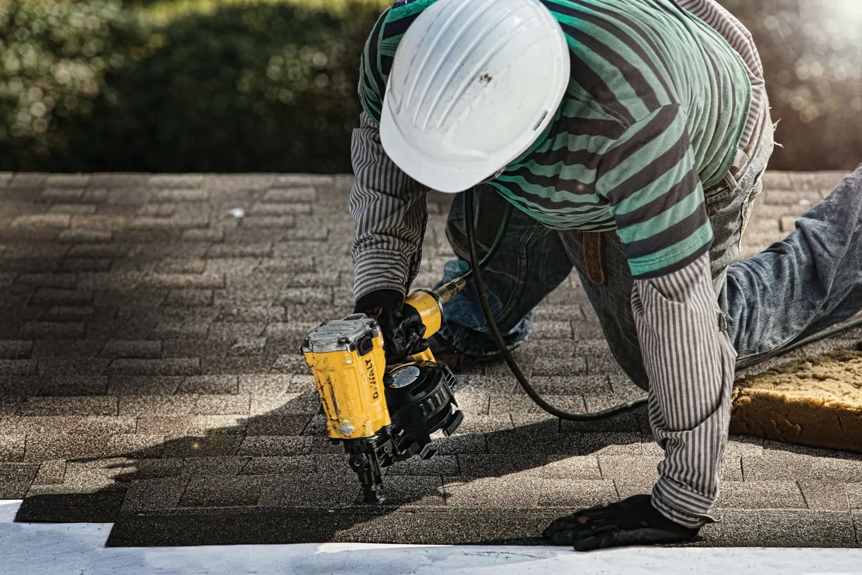 A person wearing a striped shirt, jeans, gloves, and a white safety helmet is kneeling on a roof while using a yellow DEWALT roofing nailer to install shingles.