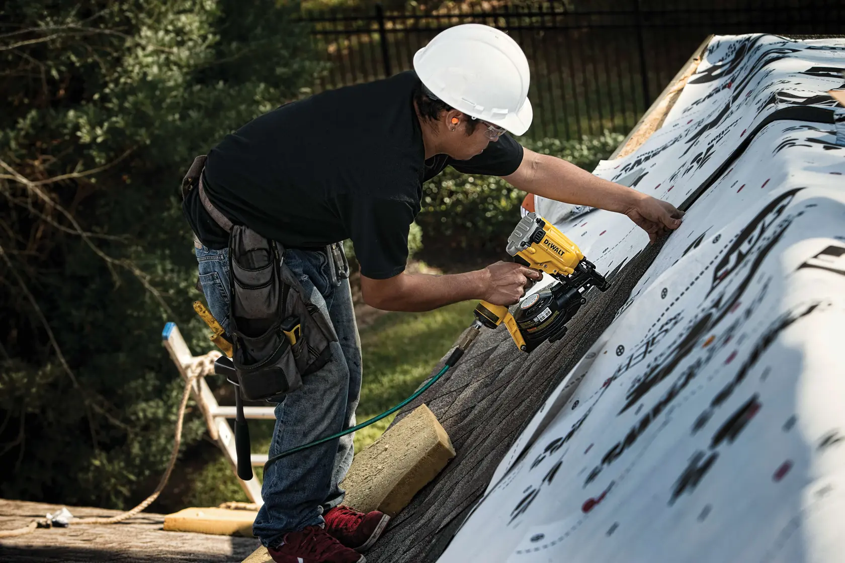 A person wearing a white safety helmet and tool belt is working on a roof, using a yellow DEWALT nail gun to secure roofing material.