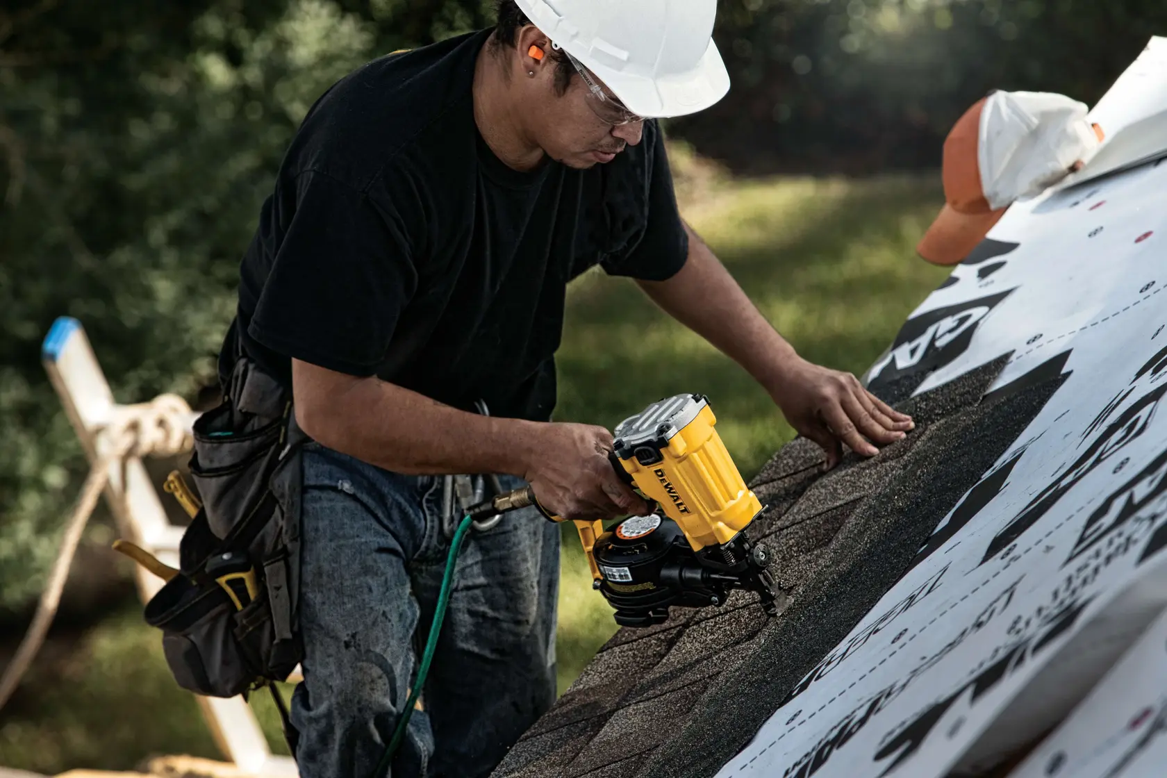A construction worker wearing a hard hat and tool belt uses a yellow DEWALT roofing nailer to install shingles on a roof.
