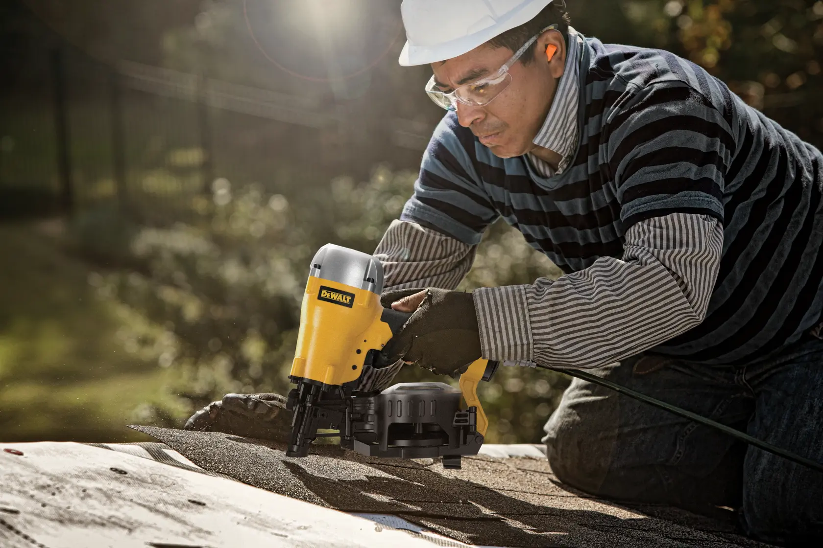 A person wearing a striped shirt, gloves, and a hard hat operates a DEWALT nail gun on a rooftop. The nail gun is yellow and is used for roofing work.