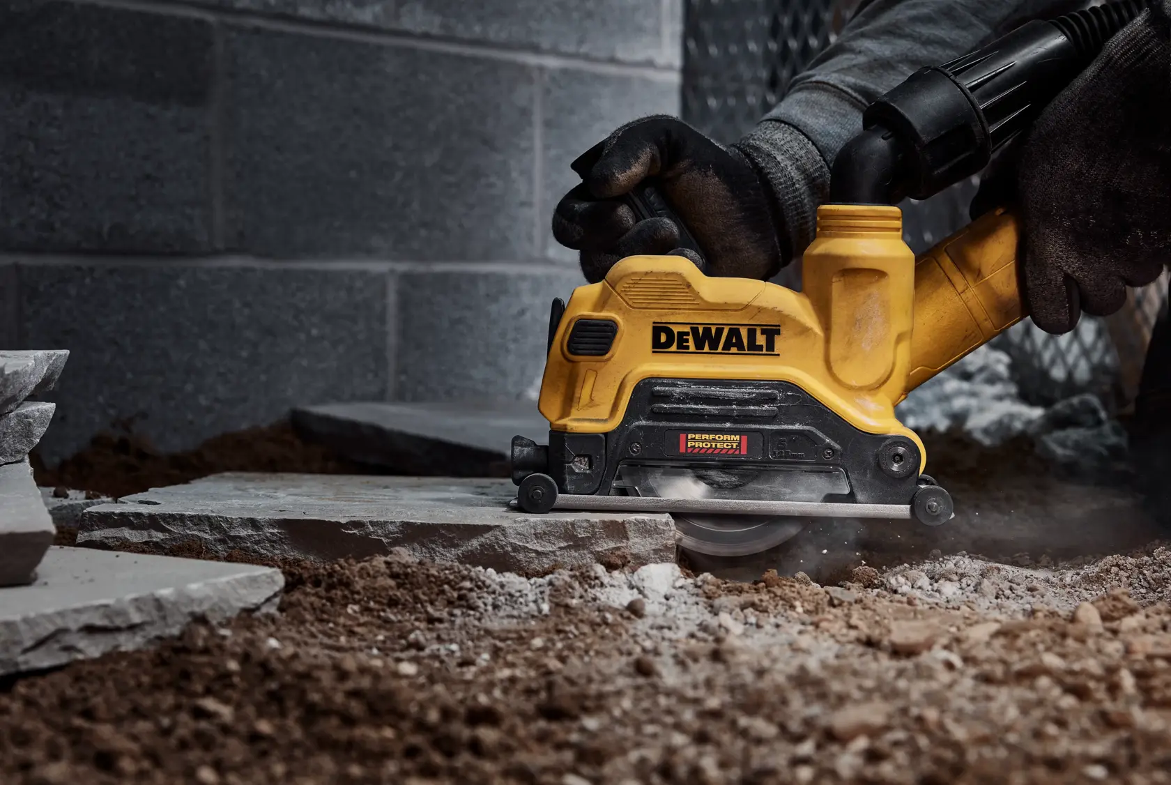 A person wearing gloves uses a DEWALT DW47227 yellow power tool to cut through stone or concrete on a construction site.
